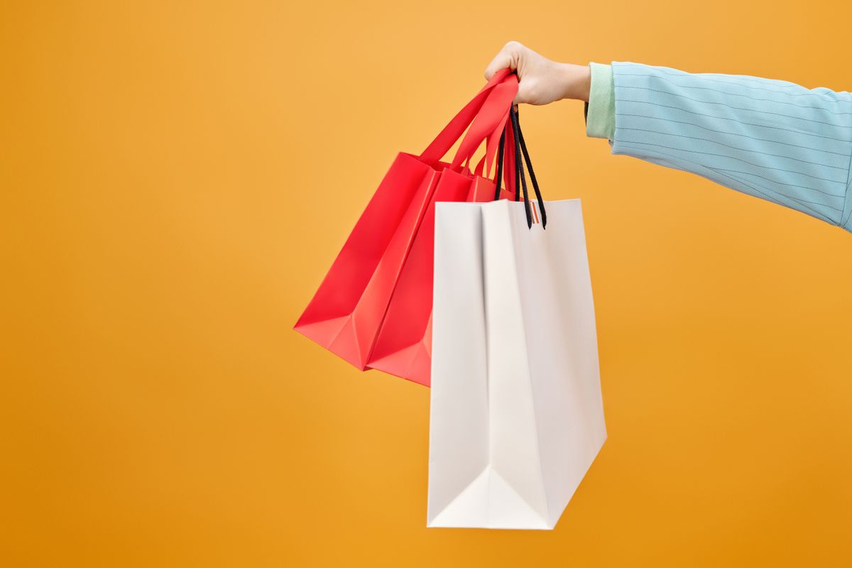 A hand holding red and white paper shopping bags against an orange background