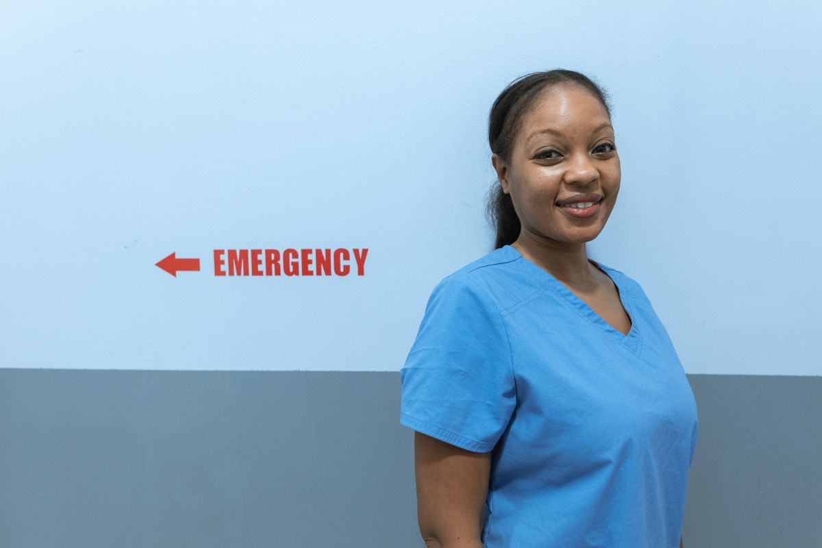 A healthcare worker in blue scrubs smiling beside a hospital emergency sign